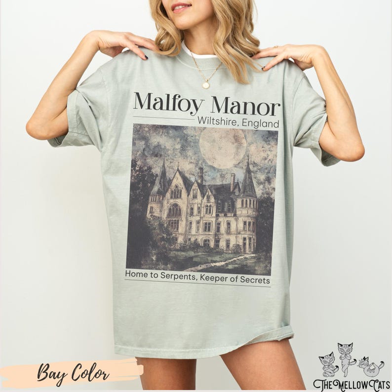 May include: A light green t-shirt featuring a graphic print of a large, grey stone manor house with a full moon in the sky. The text "Malfoy Manor" is printed above the image, and the text "Wiltshire, England" is printed below the image. The text "Home to Serpents, Keeper of Secrets" is printed below the manor house.