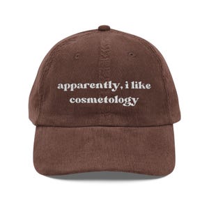 Puede incluir: Gorra de béisbol de pana marrón con el texto bordado "apparently, i like cosmetology" en blanco. La gorra tiene una visera curva y una parte superior redondeada.