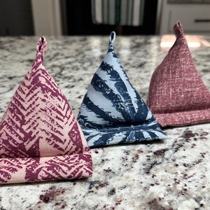 May include: Three triangular fabric phone stands in shades of burgundy, blue, and pink. The burgundy stand has a chevron pattern, the blue has a leaf design, and the pink has a textured look. Each stand has a small loop at the top.