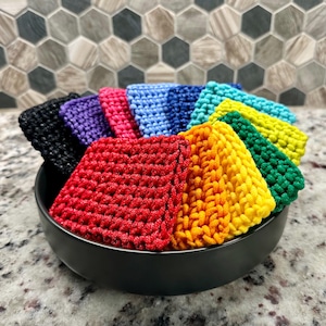 以下が含まれることがあります： さまざまな色の正方形のかぎ針編みウォッシュクロスが入った黒いボウル。ウォッシュクロスは赤、オレンジ、黄色、緑、ティール、青、紫、黒の色合いです。ボウルはまだらのカウンタートップに置かれています。