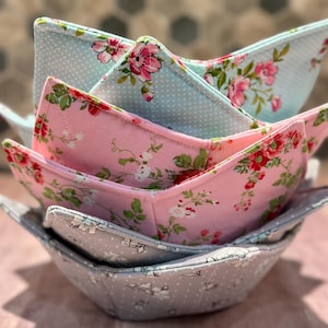 Op de afbeelding: Een stapel van vier stoffen kom-cozies in verschillende bloempatronen. De bovenste cozy is lichtblauw met witte stippen en roze bloemen. De volgende is roze met rode bloemen, gevolgd door een grijs en lichtblauw bloemmotief.