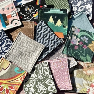 May include: A selection of square fabric coasters with diverse patterns and designs. The coasters display images of mountains, floral prints, geometric shapes, and a house with a cat. The colour palette includes green, pink, blue, and brown.