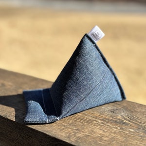 May include: A denim phone stand in a triangular shape, with a small tag that reads "MILL STREET PRODUCTS". The stand is made of blue denim fabric and is designed to hold a phone upright. The stand is placed on a wooden surface.