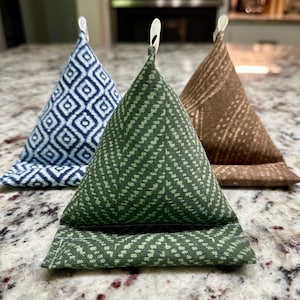 May include: Three triangular fabric phone stands in blue, green, and brown. The blue stand has a diamond pattern, the green has a chevron pattern, and the brown has a linear pattern. Each stand has a white loop at the top.
