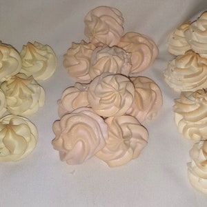 以下が含まれることがあります： メレンゲクッキーのコレクション。いくつかは白で、いくつかは薄いピンク色です。クッキーは白い表面に3つのグループに分けて配置されています。