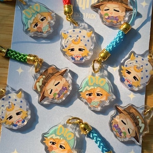 Może przedstawiać: Kolekcja akrylowych zawieszek przedstawiających postacie z anime w stylu chibi z blond włosami, noszące czapki lub nakrycia głowy, przymocowane do kolorowych plecionych sznurków ze złotymi metalowymi elementami i różnymi wyrazami twarzy.