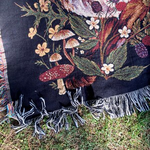 May include: Black woven throw blanket with a tapestry design of mushrooms, flowers, and leaves. The blanket has fringe on all sides.