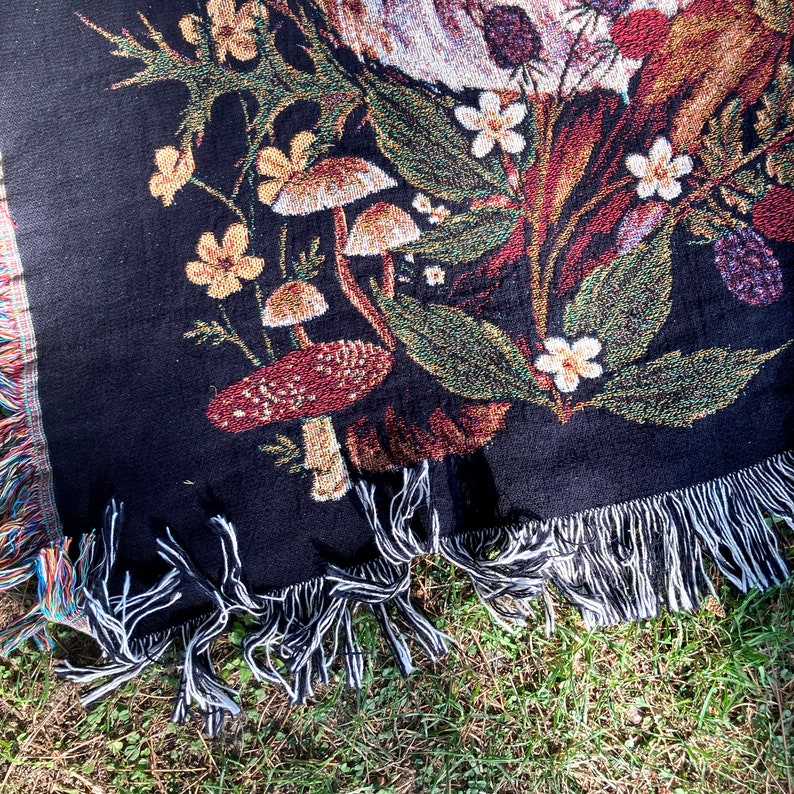 May include: Black woven throw blanket with a tapestry design of mushrooms, flowers, and leaves. The blanket has fringe on all sides.