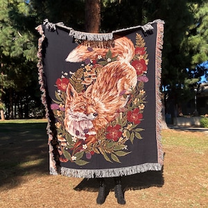 May include: A black woven throw blanket with a red fox and floral design. The fox is brown and orange with a white chest and a bushy tail. The flowers are red, pink, and white.