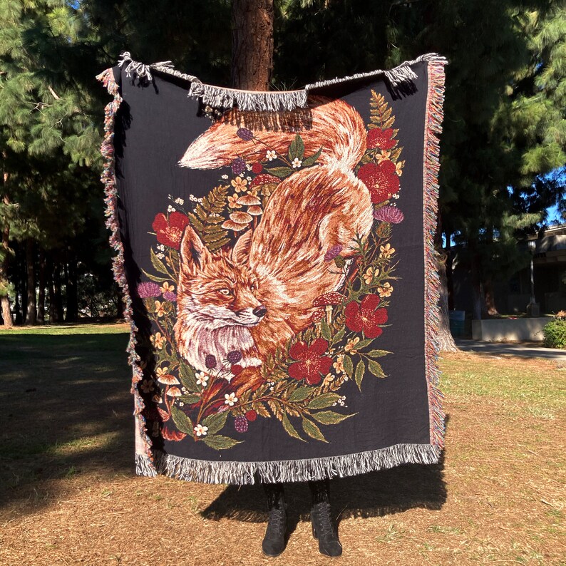 May include: A black woven throw blanket with a red fox and floral design. The fox is brown and orange with a white chest and a bushy tail. The flowers are red, pink, and white.