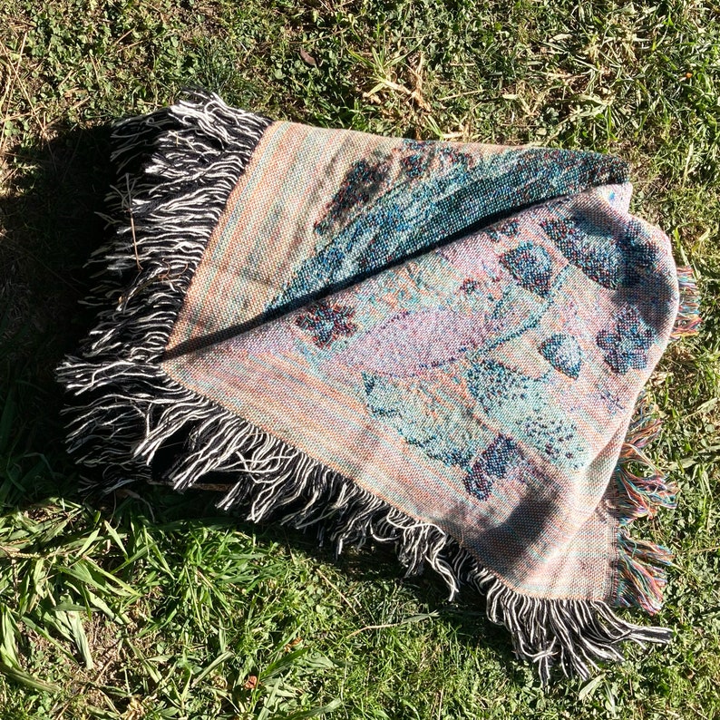 May include: A woven throw blanket with a colorful floral pattern and black fringe. The blanket is folded and lying on a bed of green grass.