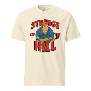 May include: Cream-colored t-shirt featuring a cartoon illustration of a person playing a guitar. The graphic includes the text "STRINGS of the HILL" in red letters, with the word "of" in a smaller font. The person in the illustration is wearing a hat and a patterned shirt.
