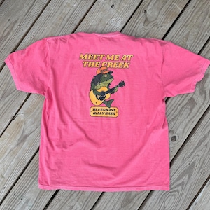 Può includere: Una maglietta rosa con la scritta "Meet Me at the Creek" e un disegno di un pesce che suona la chitarra. Il pesce indossa un cappello e la scritta "Bluegrass Billy Bass" è sotto.