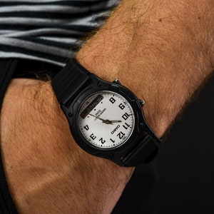 Może przedstawiać: Czarny zegarek na rękę Casio z białą tarczą i czarnymi cyframi. Zegarek ma wyświetlacz cyfrowy i sekundnik.