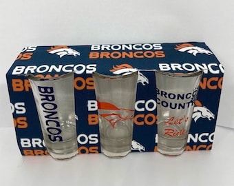 May include: Three clear shot glasses with curved tops, each featuring a different design related to the Denver Broncos. The glasses are set against a navy blue and orange Broncos-themed background. One glass has "BRONCOS" in blue, another has the team logo, and the third says "Bronco County, Let's Ride."