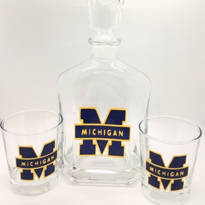 May include: A clear glass decanter set with two matching lowball glasses. Each piece features a navy blue and gold "MICHIGAN" logo. The decanter has a square base and a stopper. The glasses are cylindrical.