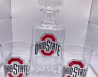 May include: A clear glass decanter with a stopper and two matching lowball glasses. Each piece features the Ohio State logo in red, gray, and black. The decanter has a square shape with a decorative pattern at the base.