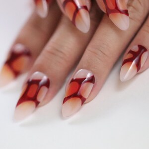 Peut inclure: Gros plan sur une main avec de longs ongles pointus. Les ongles sont peints avec un effet d&eacute;grad&eacute;, passant d'une base nude &agrave; un motif rouge vif. Le motif ressemble &agrave; une flamme ou &agrave; une forme abstraite.