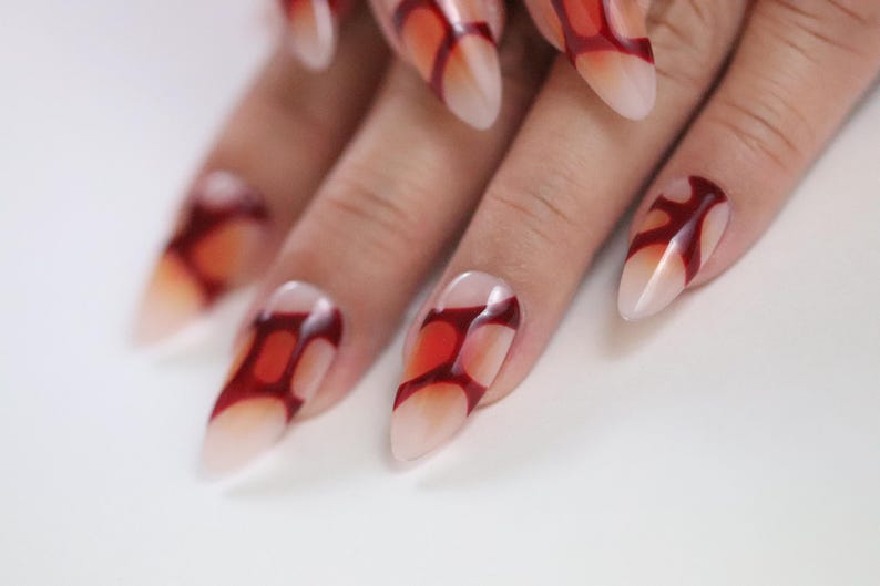 Peut inclure: Gros plan sur une main avec de longs ongles pointus. Les ongles sont peints avec un effet d&eacute;grad&eacute;, passant d'une base nude &agrave; un motif rouge vif. Le motif ressemble &agrave; une flamme ou &agrave; une forme abstraite.