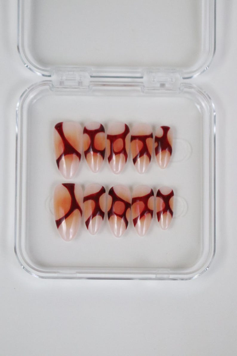 Peut inclure: Un ensemble de faux ongles ovales dans un &eacute;tui en plastique transparent. Les ongles sont rose p&acirc;le avec un motif abstrait rouge fonc&eacute;. Le motif est une s&eacute;rie de formes connect&eacute;es. L'&eacute;tui est transparent et rectangulaire.