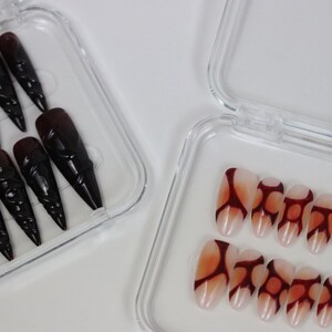 Peut inclure: Deux ensembles de faux ongles dans des bo&icirc;tes en plastique transparent. Un ensemble pr&eacute;sente de longs ongles noirs pointus avec un motif sculpt&eacute;. L'autre ensemble a une base nude avec un motif abstrait rouge fonc&eacute;. Nail art.