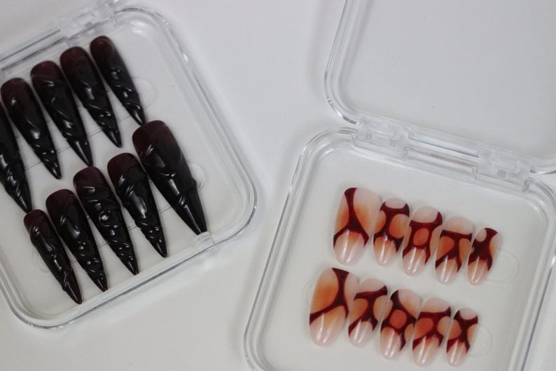 Peut inclure: Deux ensembles de faux ongles dans des bo&icirc;tes en plastique transparent. Un ensemble pr&eacute;sente de longs ongles noirs pointus avec un motif sculpt&eacute;. L'autre ensemble a une base nude avec un motif abstrait rouge fonc&eacute;. Nail art.
