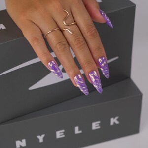 K&ouml;nnte beinhalten: Eine Hand mit langen, spitzen N&auml;geln, die in einem wirbelnden lila und goldenen Design bemalt sind. Die Hand tr&auml;gt zwei goldene Ringe, einen schlichten Bandring und einen Schlangenring. Die N&auml;gel befinden sich &uuml;ber einer grauen Box mit dem Aufdruck "NYELEK".