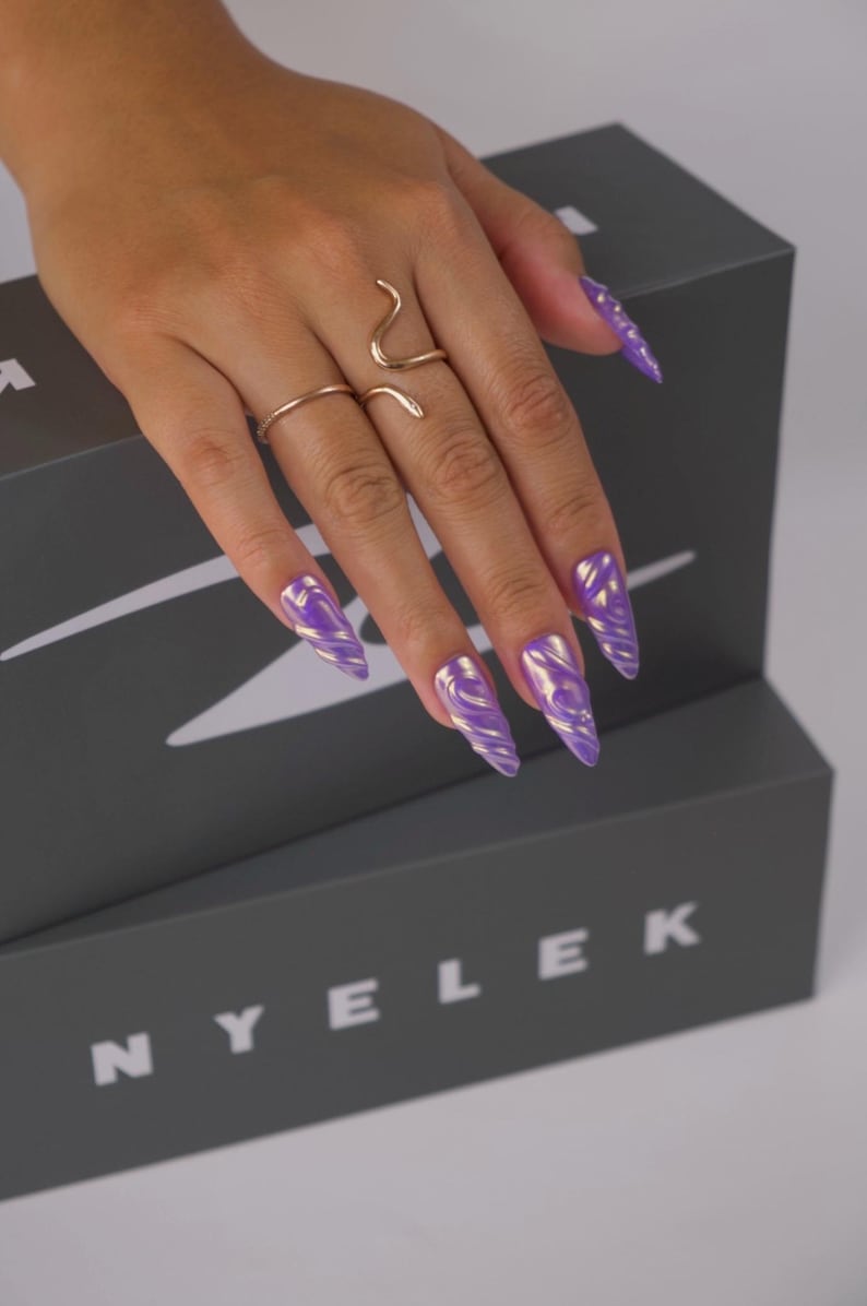 K&ouml;nnte beinhalten: Eine Hand mit langen, spitzen N&auml;geln, die in einem wirbelnden lila und goldenen Design bemalt sind. Die Hand tr&auml;gt zwei goldene Ringe, einen schlichten Bandring und einen Schlangenring. Die N&auml;gel befinden sich &uuml;ber einer grauen Box mit dem Aufdruck "NYELEK".