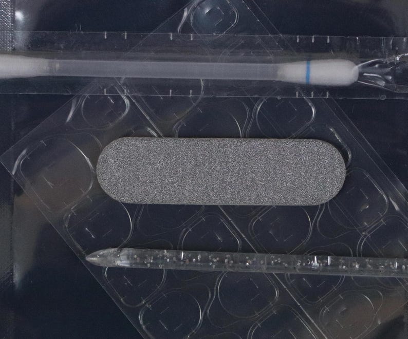 Peut inclure: Gros plan sur un emballage plastique transparent contenant une lime &agrave; ongles grise, un coton-tige et un b&acirc;tonnet en plastique transparent. La lime &agrave; ongles est rectangulaire et textur&eacute;e. Le coton-tige a une bande bleue.