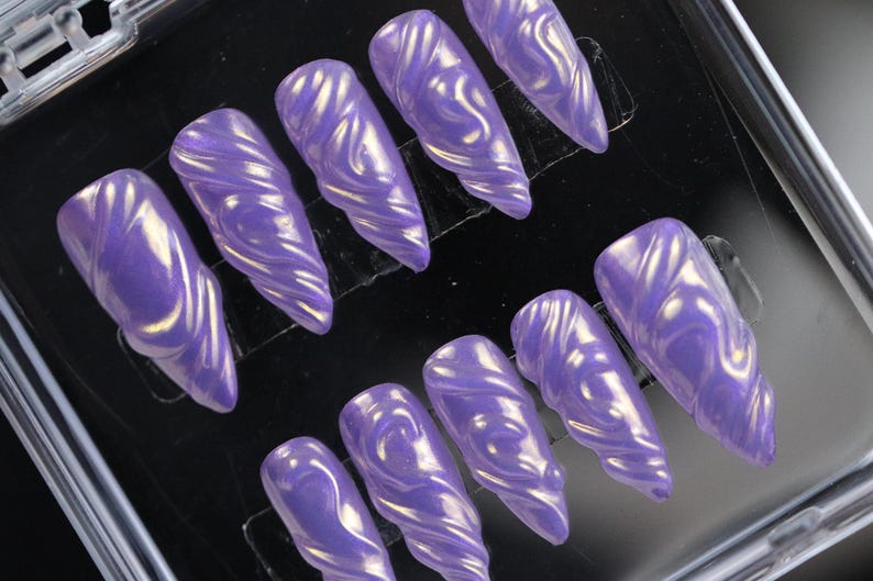 K&ouml;nnte beinhalten: Ein Set aus zehn langen, gl&auml;nzenden, lila Press-On-N&auml;geln. Jeder Nagel hat ein wirbelndes, strukturiertes Design mit goldenen Akzenten. Die N&auml;gel sind in einem transparenten Kunststoffbeh&auml;lter angeordnet.