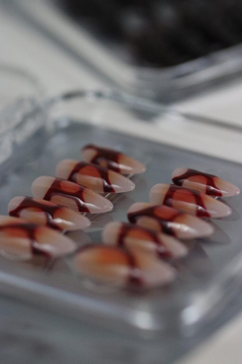 Peut inclure: Une s&eacute;rie de faux ongles en forme d'amande avec une base translucide et un motif de sang d&eacute;goulinant rouge fonc&eacute;. Les ongles sont dispos&eacute;s en rang&eacute;es sur un plateau rectangulaire transparent, cr&eacute;ant une esth&eacute;tique effrayante sur le th&egrave;me d'Halloween.