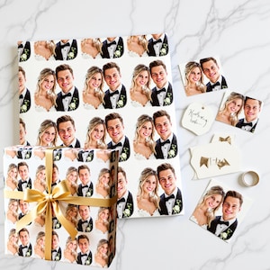 Peut inclure: Papier cadeau et étiquettes sur le thème du mariage, avec un motif répété d'un couple souriant en tenue de soirée. Une boîte cadeau emballée avec un nœud doré est à côté d'étiquettes et d'une alliance.