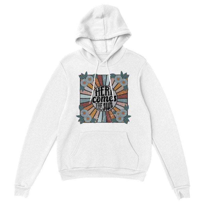 Op de afbeelding: Witte hoodie met een retro-grafische print met een zonnestralenontwerp met de tekst "Here comes the sun" in zwarte letters. De grafiek is omgeven door een rand van blauwe en gele bloemen.