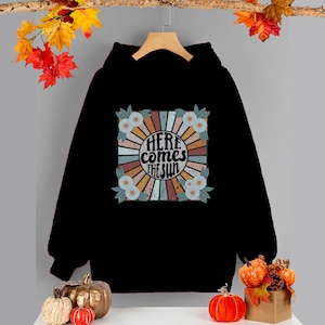 Op de afbeelding: Zwart hoodie met een retro-grafische print van een zon met de tekst "Here comes the sun" in een cirkel omringd door bloemen.