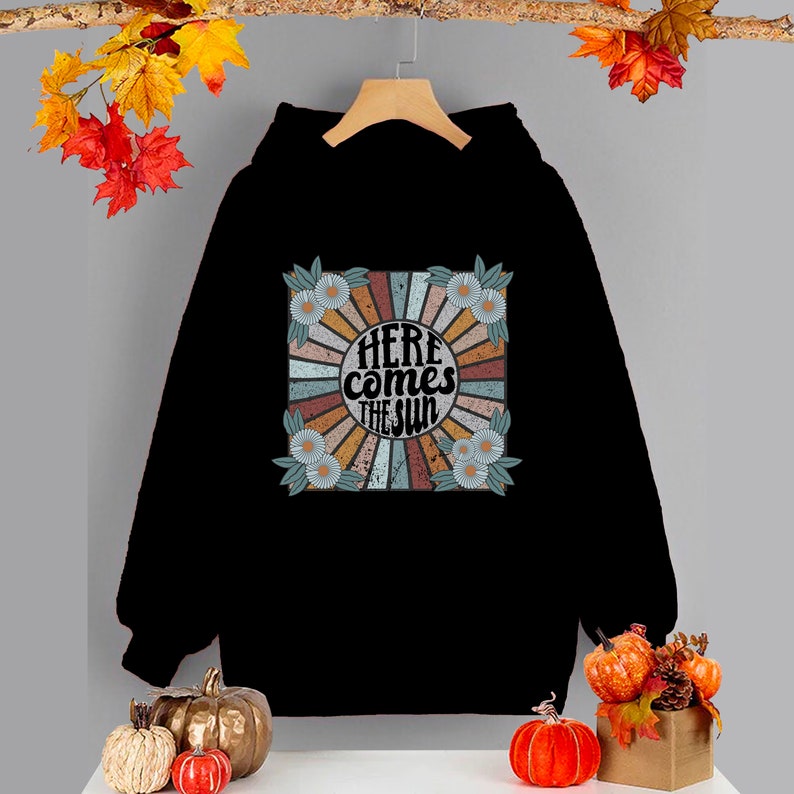 Op de afbeelding: Zwart hoodie met een retro-grafische print van een zon met de tekst "Here comes the sun" in een cirkel omringd door bloemen.
