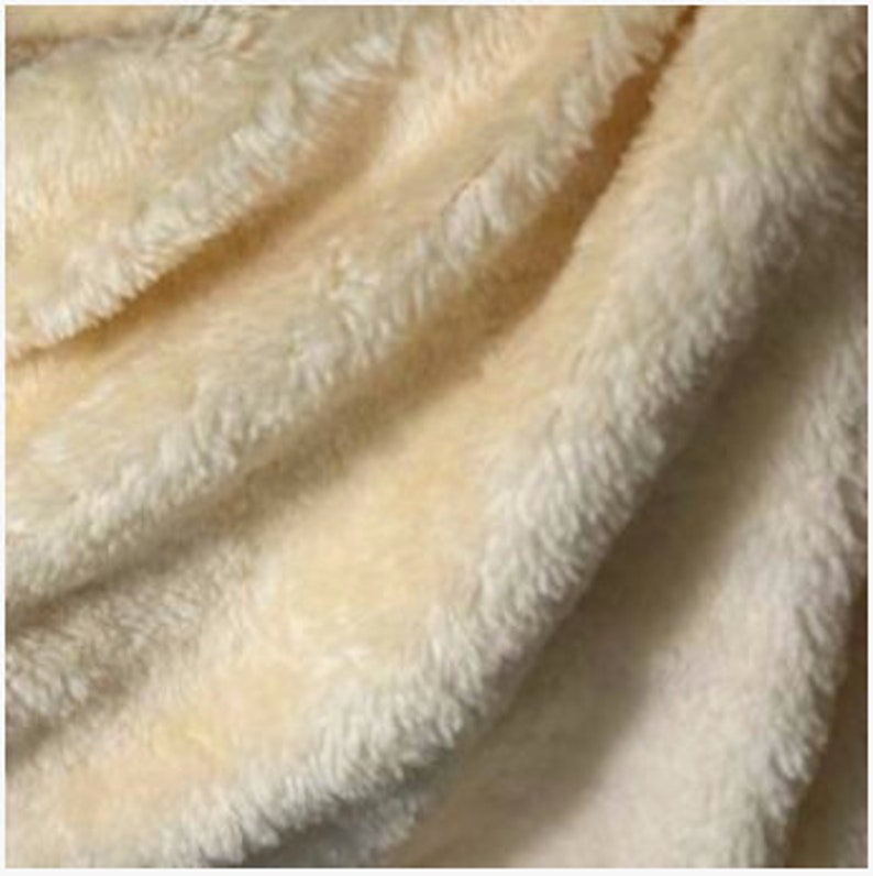 K&ouml;nnte beinhalten: Nahaufnahme einer weichen, cremefarbenen Pl&uuml;schdecke. Der Stoff hat eine zottelige Textur mit sichtbaren Fasern und Falten, die ein gem&uuml;tliches und einladendes Aussehen erzeugen. Die Decke scheint aus einem synthetischen Material zu bestehen.