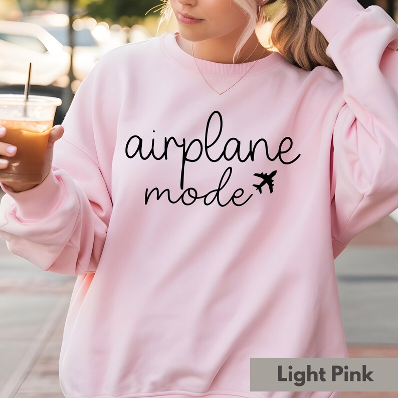 Op de afbeelding: Een lichtroze sweatshirt met de tekst "vliegtuigmodus" en een kleine vliegtuigafbeelding gedrukt op de voorkant.
