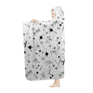 K&ouml;nnte beinhalten: Eine wei&szlig;e Kapuzen-Decke mit einem schwarz-wei&szlig;en Weltraumschlacht-Design. Die Decke hat ein sich wiederholendes Muster aus Raumschiffen, Sternen und Explosionen. Die Person h&auml;lt die Decke.