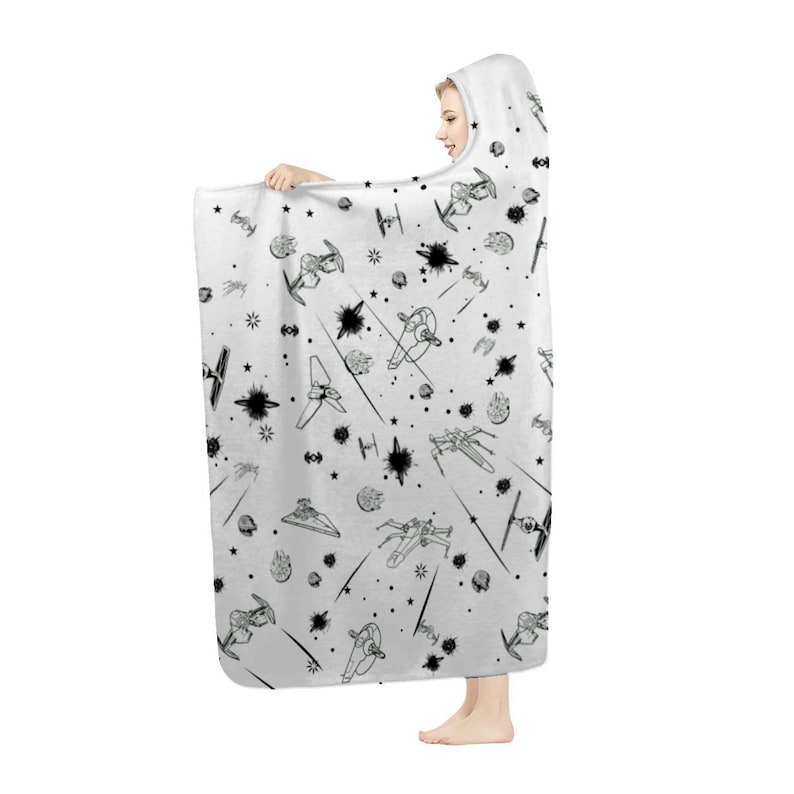 K&ouml;nnte beinhalten: Eine wei&szlig;e Kapuzen-Decke mit einem schwarz-wei&szlig;en Weltraumschlacht-Design. Die Decke hat ein sich wiederholendes Muster aus Raumschiffen, Sternen und Explosionen. Die Person h&auml;lt die Decke.