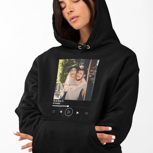 May include: Black hoodie with a white graphic of a couple in a photo frame with a music player interface below. The music player interface shows the song title "Te la Doy" by "El Ankla" and play, pause, rewind, and fast forward buttons.