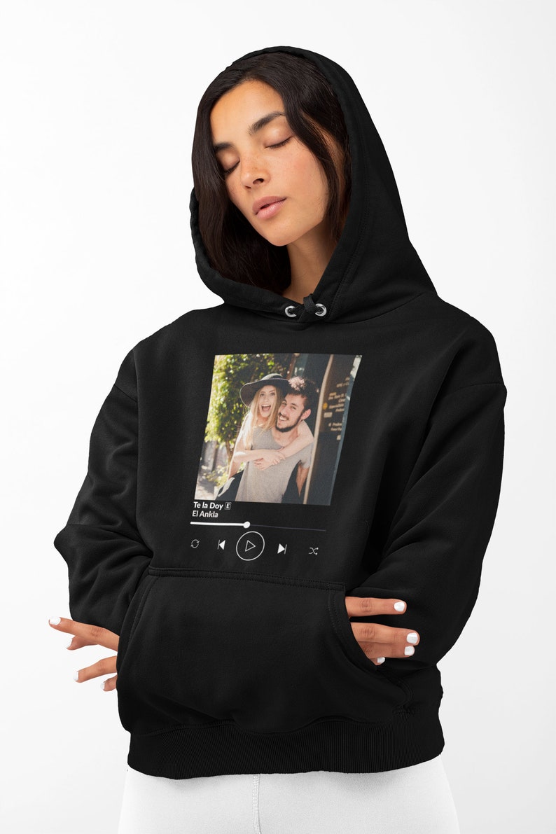 May include: Black hoodie with a white graphic of a couple in a photo frame with a music player interface below. The music player interface shows the song title "Te la Doy" by "El Ankla" and play, pause, rewind, and fast forward buttons.