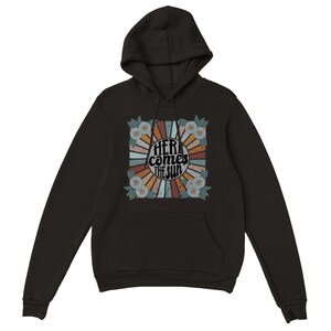 Op de afbeelding: Zwart hoodie met een retro-grafische print met een zonnestralenontwerp met de tekst "Here comes the sun" in een cirkel omringd door bloemen.
