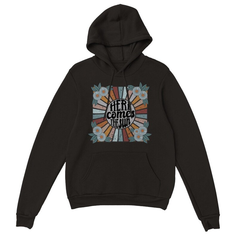 Op de afbeelding: Zwart hoodie met een retro-grafische print met een zonnestralenontwerp met de tekst "Here comes the sun" in een cirkel omringd door bloemen.