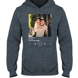 May include: A dark gray hoodie with a photo of a couple on the front. The photo is in a white frame with a music player interface below it. The music player interface shows the song title "Te la Doy" by "El Ankla" and play, rewind, and fast forward buttons.