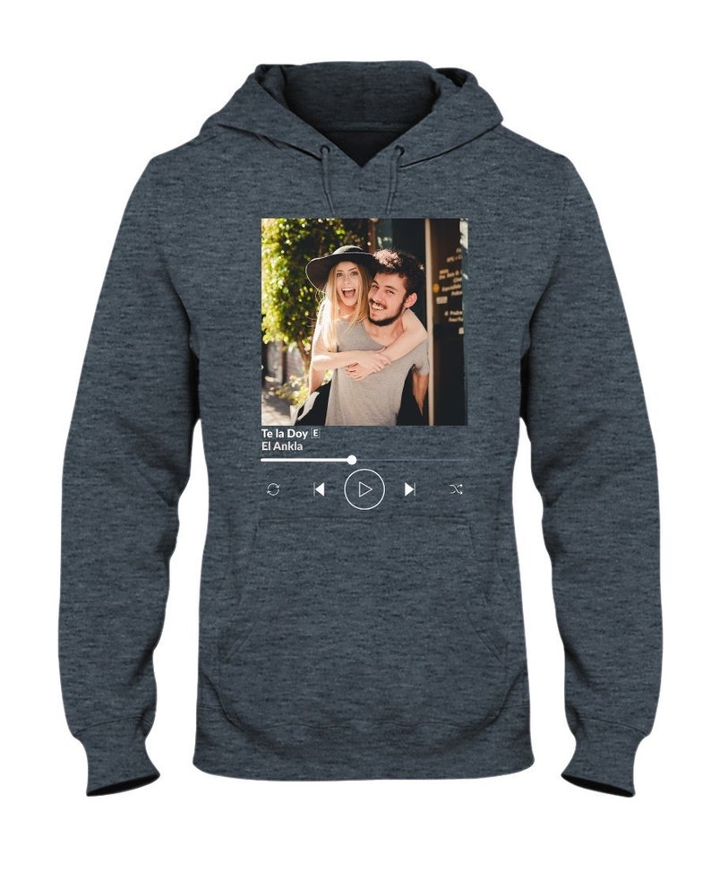 May include: A dark gray hoodie with a photo of a couple on the front. The photo is in a white frame with a music player interface below it. The music player interface shows the song title "Te la Doy" by "El Ankla" and play, rewind, and fast forward buttons.