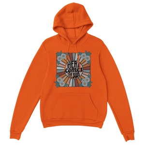 Op de afbeelding: Oranje hoodie met een retro-grafische print met een zonnestralenontwerp en de tekst "Here comes the sun" in een vintage lettertype.