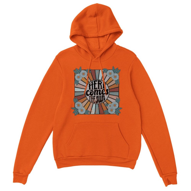 Op de afbeelding: Oranje hoodie met een retro-grafische print met een zonnestralenontwerp en de tekst "Here comes the sun" in een vintage lettertype.