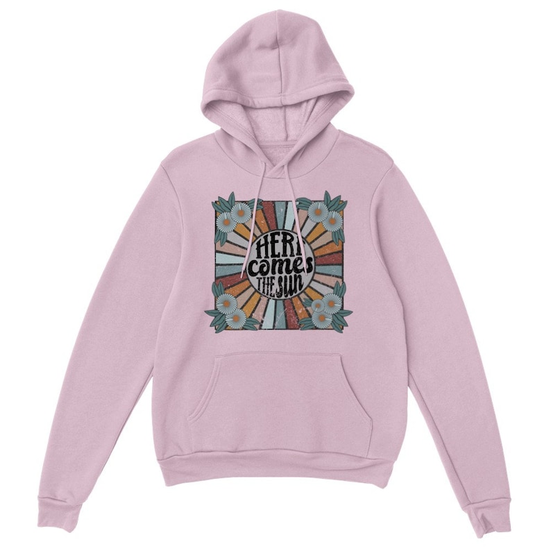 Op de afbeelding: Een roze hoodie met een grafische print van een retro zonnestralenontwerp met de tekst "Here comes the sun" in zwart. De grafiek is omgeven door een bloemige rand.