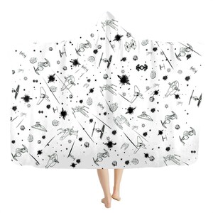 K&ouml;nnte beinhalten: Wei&szlig;e Kapuzen-Decke mit einem schwarz-wei&szlig;en Star Wars-Muster. Das Design zeigt Raumschiffe, Explosionen und Sterne. Die Decke ist &uuml;ber einer Person drapiert.