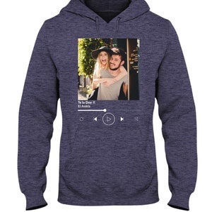 May include: A navy blue hoodie with a photo of a couple on the front. The photo is in a white frame with a music player interface below it. The music player interface shows the song title "Te la Doy" by "El Ankla" and play, rewind, and fast forward buttons.